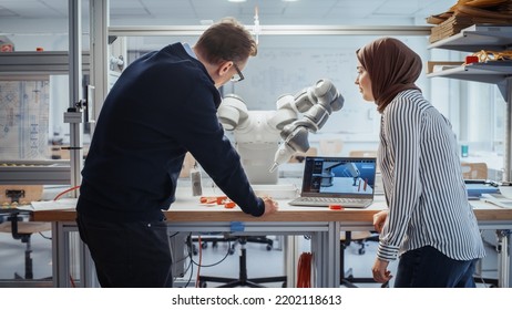 Senior Male Engineer Explaining Controls To Muslim Female Computer Science Specialist And Pointing At Robotic Hand In The Research Laboratory With Laptop. Innovative Startup Company Concept.