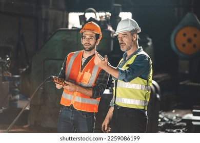 Senior male engineer training and explaining work to new employee wearing vest and safety jacket with hardhat helmet while pointing towards machine in factory and giving instructions - Powered by Shutterstock