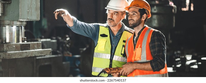 Senior male engineer training and explaining work to new employee wearing vest and safety jacket with hardhat helmet while pointing towards machine in factory and giving instructions - Powered by Shutterstock