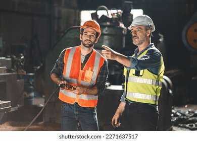 Senior male engineer training and explaining work to new employee wearing vest and safety jacket with hardhat helmet while pointing towards machine in factory and giving instructions - Powered by Shutterstock