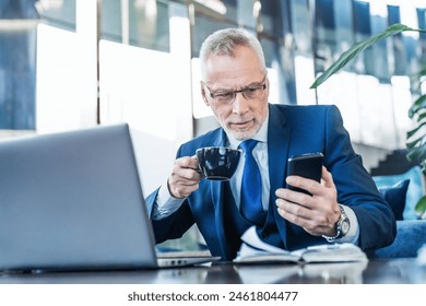 Senior male executive drinking coffee and holding smartphone while working online in office lounge. Business manager ceo using cell phone mobile app, laptop, computer