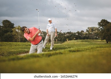 Senior male golfer playing golf on the course. Man hitting the ball out of a sand bunker with his opponent standing at the back. - Powered by Shutterstock