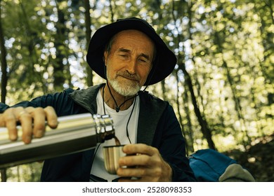 Senior male in hat pouring coffee from a thermos while taking a break in a forest during walk - Powered by Shutterstock