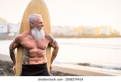 Senior Male Having Fun Surfing During Sunset Time - Fit Retired Man Training With Surfboard On The Beach - Elderly Healthy People Lifestyle And Extreme Sport Concept