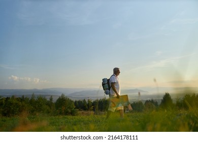 Senior Male Hiker Walking In Green Valley, While Hiking In Dusk. Side View Of Adult Traveler Holding Map, While Backpacking In Mountains, With Scenic Landscape On Background. Concept Of Backpacking.