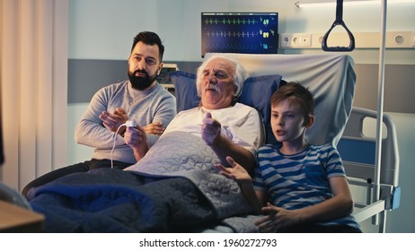 Senior male patient discussing match with adult man and boy then celebrating victory while lying on bed and watching TV in ward of contemporary clinic