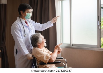 Senior Male Patient Sitting On Wheelchair In Hospital Smiling And Talking And Showing Something Outside The Window To Handsome Doctor In Labcoat In Room