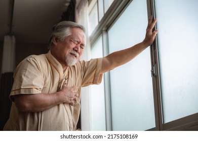 Senior Male Patient Wearing Gown Admitted In Hospital Holding Chest Due To Pain Suffering From Heart Attack While Holding And Taking Support Of Window In Ward And Private Room