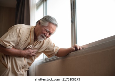 Senior Male Patient Wearing Gown Admitted In Hospital Holding Chest Due To Pain Suffering From Heart Attack While Holding And Taking Support Of Window In Ward And Private Room