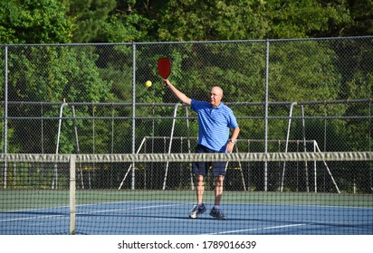 Senior Male Returns A High Hit While Playing Pickle Ball, On An Outside Tennis Court.  He Has On A Blue Striped Shirt.