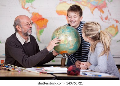 Senior male teacher pointing at globe while students looking at it at desk in classroom - Powered by Shutterstock