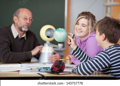 Senior Male Teacher And Students Looking At Planetarium Model At Desk In Classroom