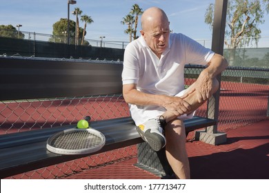 Senior Male Tennis Player With Leg Pain Sitting On Bench At Court