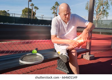 Senior Male Tennis Player With Leg Pain Sitting On Bench At Court