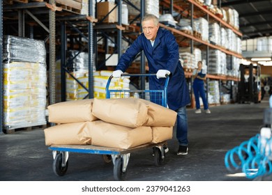 Senior male worker carries, pushes, cart for cargo transportation. Elderly man works as loader in rack area warehouse of construction materials - Powered by Shutterstock