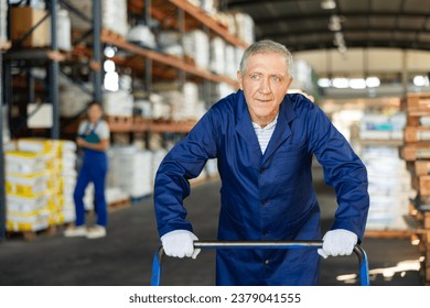 Senior male worker carries, pushes, cart for cargo transportation. Elderly man works as loader in rack area warehouse of construction materials - Powered by Shutterstock