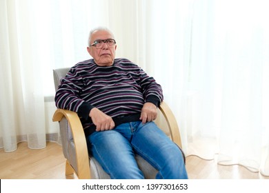 Senior Man 70-75 Years Old Sitting In An Armchair And Looking At Camera. On Background Big Light Window. On Right Side Have Space For Text Ad