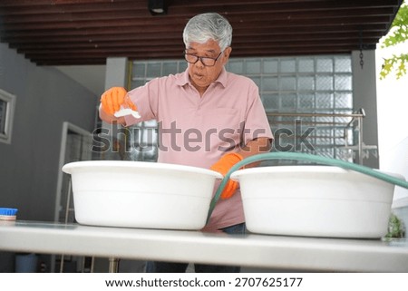 Senior man adding detergent while doing laundry outdoors, Elderly man preparing wash water for cleaning clothes, Older adult working on household laundry chores outside