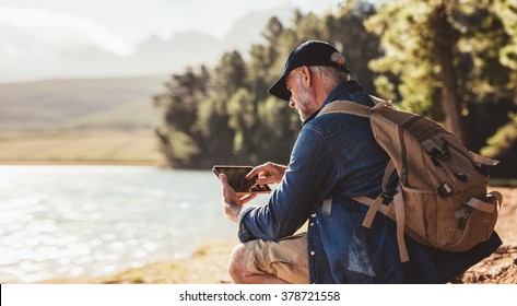 Senior man with backpack using digital tab while sitting near a lake. Mature man on hike in nature using digital tablet. - Powered by Shutterstock