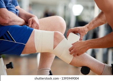 Senior Man With Bandage On Leg After Injury In Gym. Male Hands Fixed Elastic Bandage On Male Injured Leg. Sport And Health.