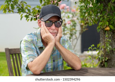 Senior Man In A Baseball Cap With Sunglasses Relaxing In The Garden