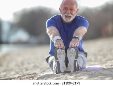 Senior man with beard in sportswear stretching on the beach while sitting in the sand and wearing wristband and smart watch selective focus - Powered by Shutterstock