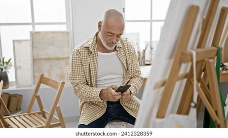 A senior man with a beard uses a smartphone in an art studio filled with easels and paintings. - Powered by Shutterstock