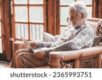 Senior man with beard and white hair sitting comfortably at home on armchair using laptop. Older relaxed person and authentic retirement lifestyle