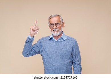 Senior man in black-rimmed glasses points index finger up showing hand gesture and smiling. Cheerful elderly bearded person businessman stands at beige wall