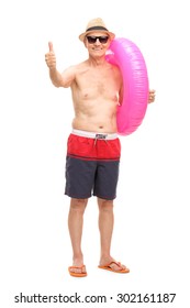 Senior Man In Blue Swim Trunks Carrying A Big Pink Swimming Ring And Giving A Thumb Up Isolated On White Background