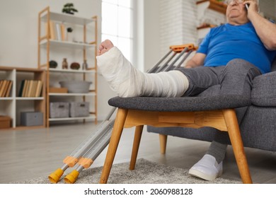 Hombre mayor con la pierna rota en yeso de yeso de yeso sentado en el sofá de casa y hablando por teléfono móvil. Paciente de edad avanzada con pie herido descansando en el sofá y llamando al doctor por teléfono celular. Cerrar