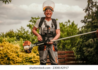 Senior man with brush cutter on the lawn against the background of bushes in workwear and goggles