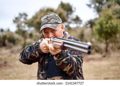 Senior Man In Camouflage Clothes And Cap Squinting And Aiming Shotgun While Hunting In Countryside On Autumn Day