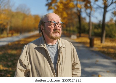 Senior man in casual attire walks outdoors in a park during the autumn season, with fallen leaves and colorful foliage creating a serene, natural setting. - Powered by Shutterstock