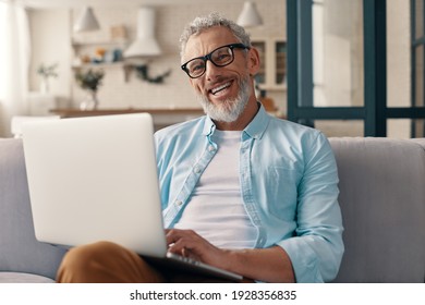 Senior man in casual clothing using laptop and looking at camera with smile while sitting on the sofa at home - Powered by Shutterstock