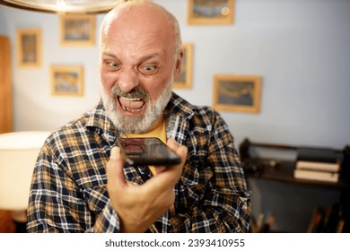 Senior man in checkered shirt yelling at smartphone. Angry furious bald bearded man recording voice message, expressing rage and displeasure, shouting with opened mouth, scolding someone - Powered by Shutterstock