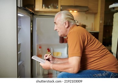 Senior Man Checking His Fridge And Making Shopping List Before Going To Grocery Store