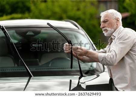 Senior man checking and replacing the windshield wiper blade on his car, ensuring safe visibility for summer road trips