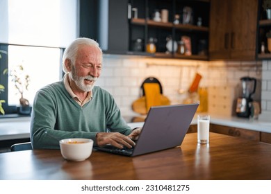 Senior man connected on internet with laptop at home. Senior man with cup of tea using laptop at table in kitchen. Mature man typing on laptop in kitchen during breakfast and driking coffee. - Shutterstock ID 2310481275
