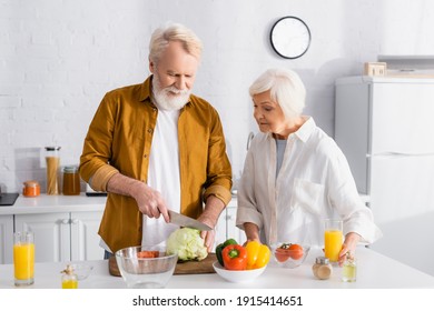 Senior man cutting cabbage near vegetables and wife with orange juice - Powered by Shutterstock