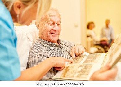 Senior Man With Dementia Looks At Photos Together With Caregiver In Nursing Home