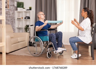 Senior Man With Disability In Wheelchair Doing Recovery Exercise With Resistance Band. Disabled Handicapped Old Person With Social Worker In Recovery Support Therapy Physiotherapy Healthcare System