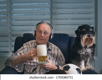 Senior Man With Dog Sitting In Armchair At Home, Holding Soccer Ball And Mug Of Beer And Watching Tv