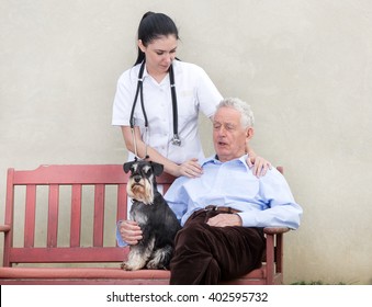 Senior Man With Dog Sitting On The Bench While Young Nurse Standing Behind Him And Holding Her Hand On His Shoulder