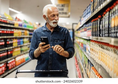 Senior Man doing grocery shopping at the supermarket, he is using apps on his smartphone. Male customer shopping with smartphone checklist, taking products from shelf at the shop. - Powered by Shutterstock