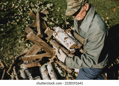 Senior man dressed in jacket and cap holding firewood in his hands. Old man preparing for winter, caring heap of wood logs. Heating with wood and solid fuel. Energy saving concept - Powered by Shutterstock