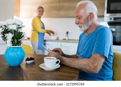 Senior man drinking coffee and looking at tablet at dining  table while woman preparing food in kitchen behind him - Powered by Shutterstock