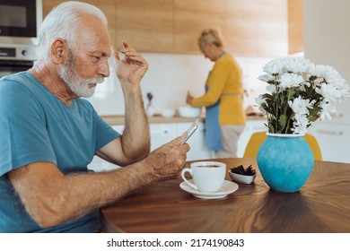 Senior man drinking coffee and looking at mobile phone at dining  table while woman preparing food in kitchen behind him - Powered by Shutterstock