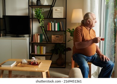 Senior Man Drinking Cup Of Black Tea At Home And Looking Outside Through Big Window