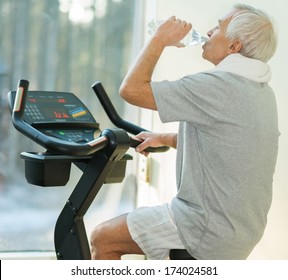 Senior Man Drinking While Doing Exercise On A Bike In A Fitness Club